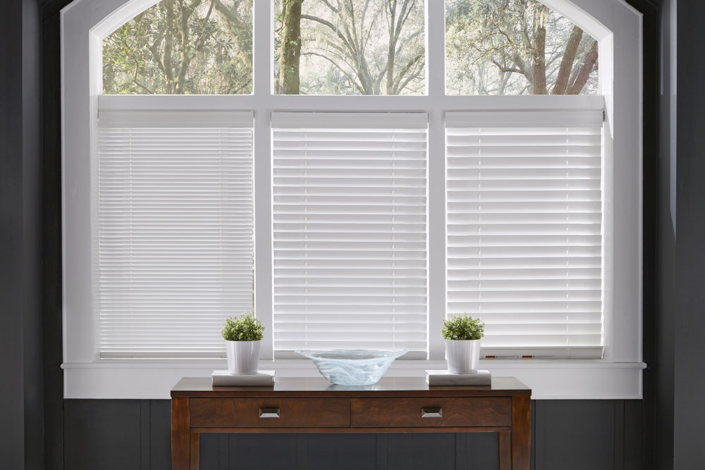 three white Heartland Woods® Wood Blinds in 1", 2" and 2 1/2" slat sizes in the open position behind a brown desk against dark gray walls