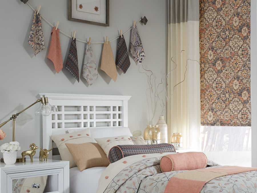 closeup of a bed with pink, purple, light pink, tan, and white Interior Masterpieces® Pillows and matching Custom Bedding with a window that has Draperies and Fabric Shades that match
