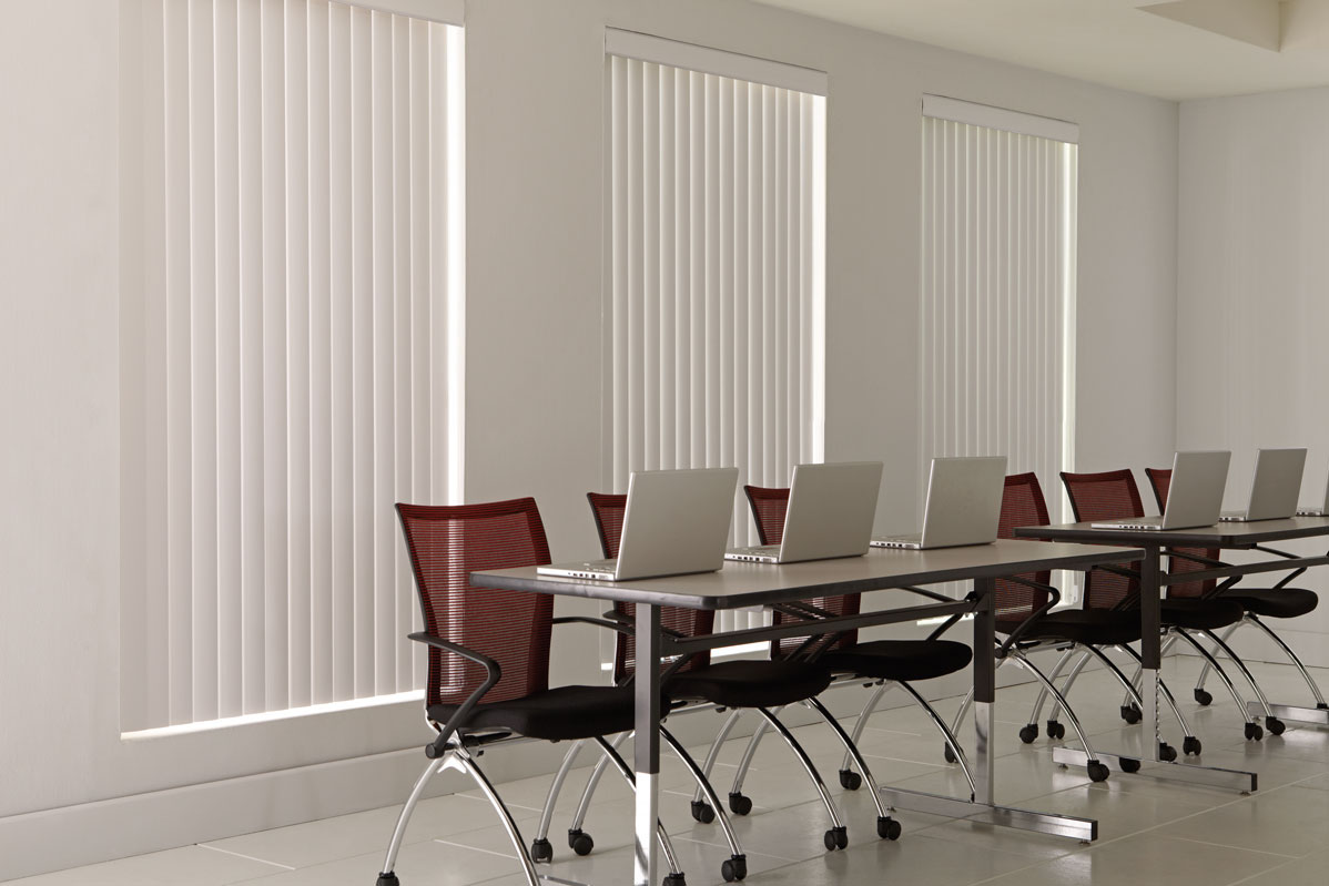 3 large windows with white colored Discoveries® Vertical Blinds in them in an office with several red chairs and laptops