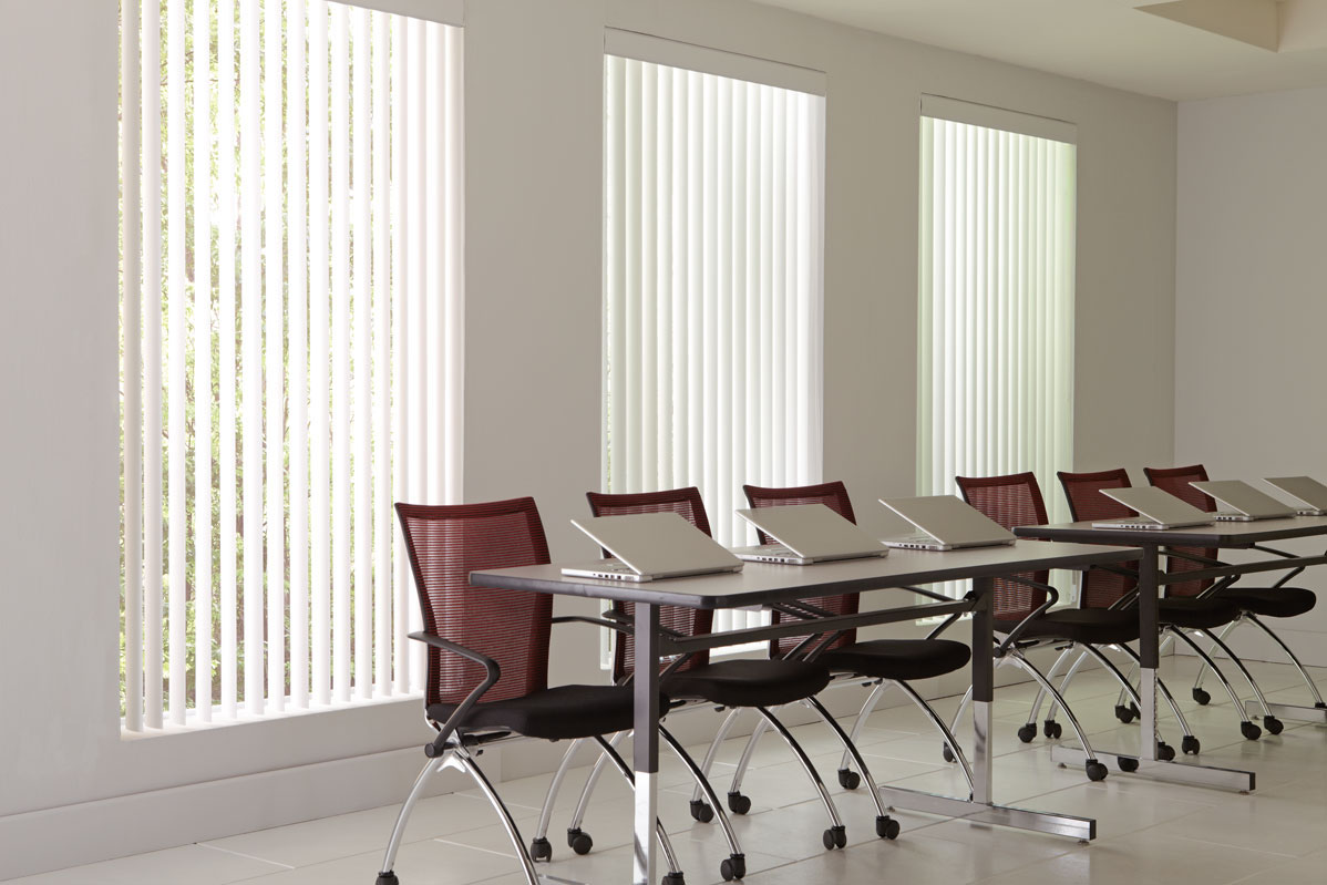 3 large windows with white colored Discoveries® Vertical Blinds in them in an office with several red chairs and laptops
