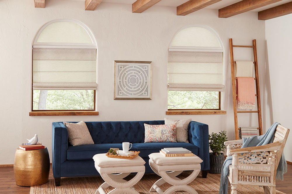 Tan colored Genesis® Hobbled Roman Shades with half circle Custom Shapes and a Palladian Shelf between behind a blue couch with tan pillows