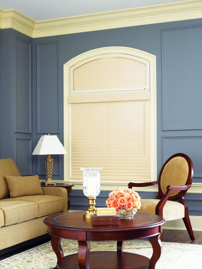 a cream colored Heartland Woods® Wood Blind with a specialty curved top shape above against a blue wall with a tan couch and mahogany table and chair in front