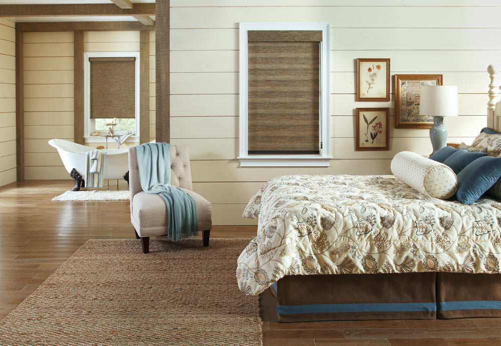 Brown Allure® Transitional Shade behind a bed with Interior Masterpieces® Custom Pillows and Bedding across from a room with a brown Genesis® Roller Shade and Panel Track