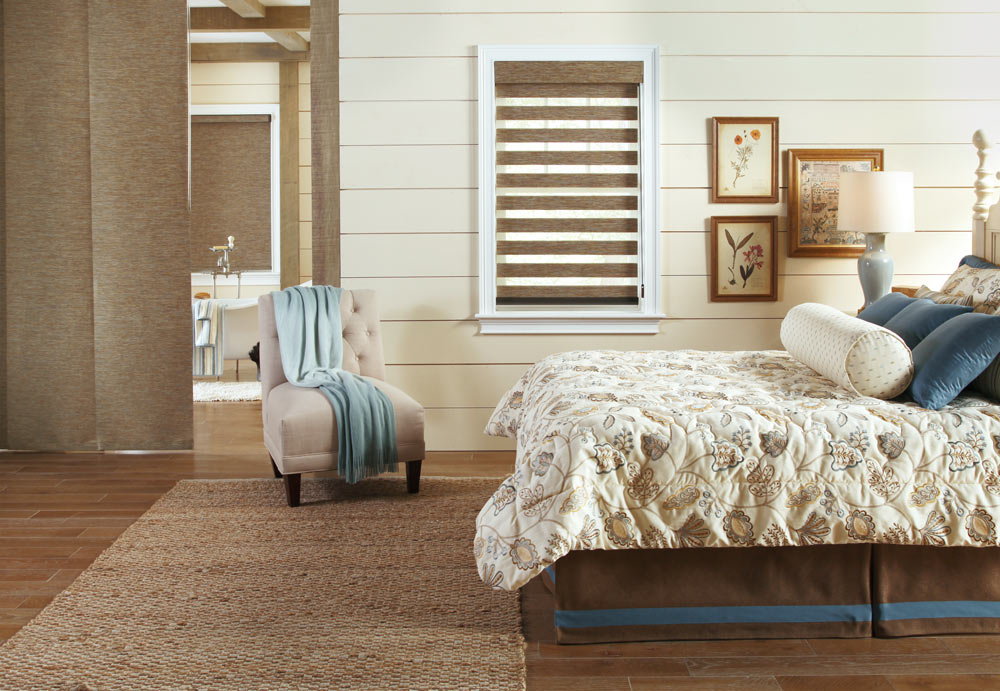 Brown Allure® Transitional Shade behind a bed with Interior Masterpieces® Custom Pillows and Bedding across from a room with a brown Genesis® Roller Shade and Panel Track